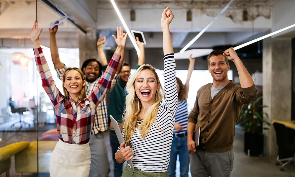Blog - Professionals Smiling and Celebrating in their Work Office Halway While Holding Laptops and Tablets and Their Arms in the Air
